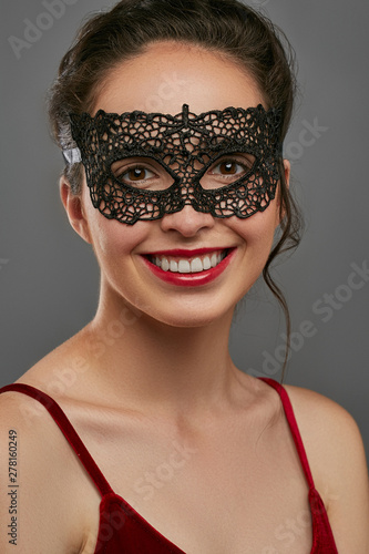 Portrait of smiling woman with tied back dark hair, wearing wine red crop top. The young girl is looking at camera, wearing black carnival mask with perforation. Vintage women's carnival accessory.