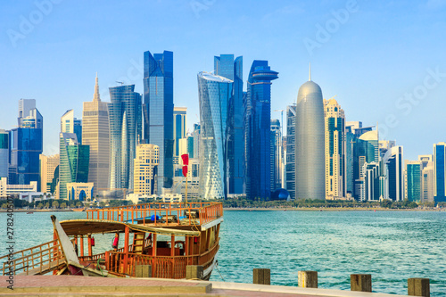 Traditional wooden dhow on foreground at Doha Bay and skyscrapers towers of West Bay skyline on background. Capital of Qatar, Middle East, Persian Gulf. Sunny blue sky. Urban modern cityscape.
