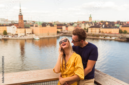 Portrait of the cheerful young couple who is standing in the embracing against the background of the city and river and a woman speaking by phone