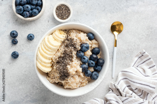 Breakfast oatmeal porridge with banana, blueberries, chia seeds on grey concrete background. Table top view of healthy food for weight loss, clean eating, vegan and vegetarian diet