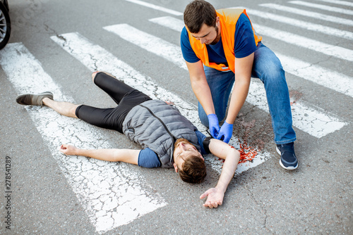 Man applying first aid to the injured bleeding person, wearing tourniquet on the arm after the road accident on the pedestrian crossing