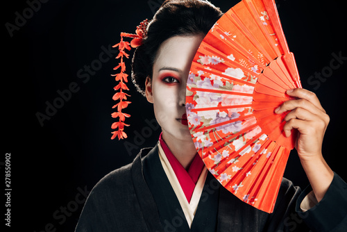 attractive geisha in black kimono with flowers in hair holding bright hand fan isolated on black