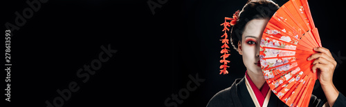 attractive geisha in black kimono with flowers in hair holding bright hand fan isolated on black, panoramic shot