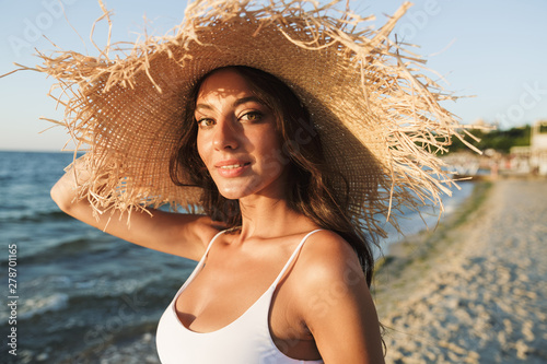 Photo of caucasian brunette woman in swimsuit and straw hat smiling and sunbathing while walking by seaside