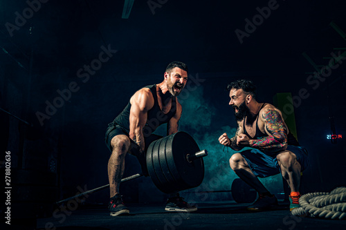 Two muscular bearded tattoed athletes training, one lift heavy weight bar when other is motivating. Scream. Working hard. Exercise for the muscles of the back