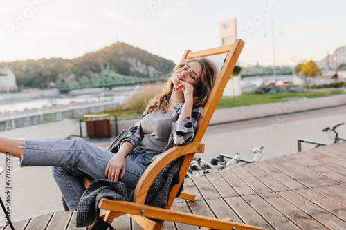 Dreamy european girl in jeans sleeping in recliner on amazing nature background. Stunning white lady in casual clothes chilling in park chaise-longue morning.