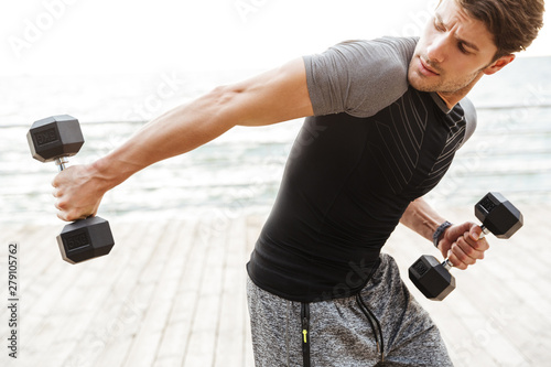 Strong handsome concentrated young sports man outdoors at beach make exercises for triceps arms with dumbbells.