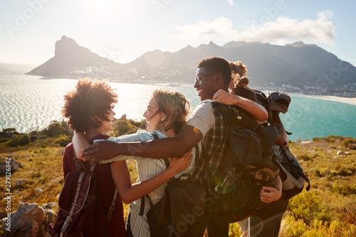 Millennial friends on a hiking trip embrace at the summit to celebrate their climb, back view