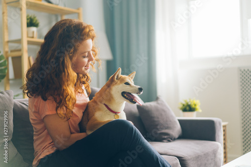 Pretty redhead woman is hugging cute doggy sitting on couch in apartment smiling enjoying beautiful day with beloved animal. People and pets concept.