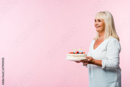 happy retired woman with grey hair holding tasty birthday cake isolated on pink