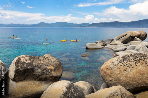 Kayakers and paddleboarders paddling on the turquoise crystal-clear water of Sand Harbor in Lake Tahoe with giant boulders in the foreground and mountains in the background
