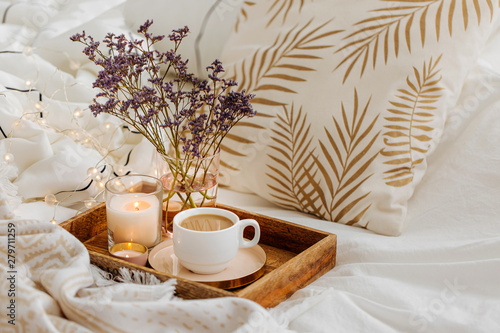 Wooden tray of coffee and candles with flowers on bed. White bedding sheets with striped blanket and pillow. Breakfast in bed. Hygge concept.