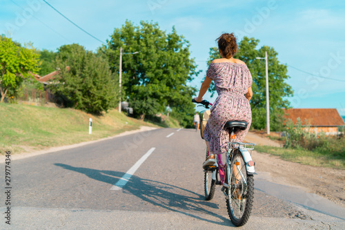 Back view of Young woman in a sunny summer day wearing dress riding a bike bicycle on the open road trough the village