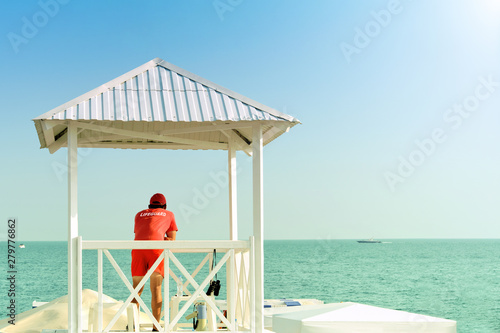 lifeguard on sea beach in watch tower on rescue duty against blue ocean water background back view of person in red t-shirt and swimming shorts with lifeguard label guarding sea shore outdoor activity