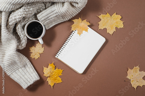 Workspace with golden maple leaves, notebook, coffee cup, sweater on brown background. Creative composition. Autumn or Winter concept. Flat lay, top view