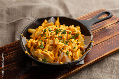 Mouthwatering hot french fries with a lot of cheese sauce with herbs in a pan, on a wooden board