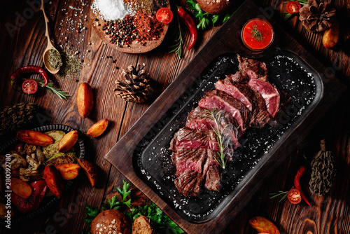 Juicy roasted veal tenderloin sliced decorated rosemary on dark board on dark wooden background in beautiful composition among vegetables and spices. Top view. Flat lay