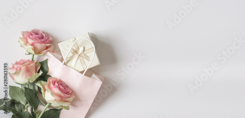 Top view composition with pastel pink three roses, paper bag and light gift box with a bow on a white background. Romantic valetine, womens day, spring sale banner copy space.