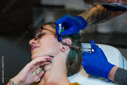 Portrait of a woman getting her ear pierced. Man showing a process of piercing ear with steril neadle and latex gloves. Ear Piercing Procedure