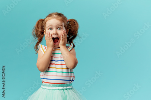 Portrait of surprised cute little toddler girl child standing isolated over blue background. Looking at camera. hands near open mouth