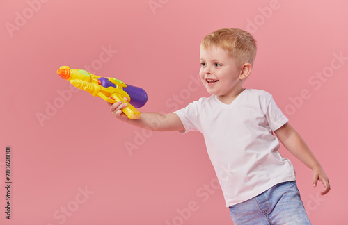 Happy little toddler boy in summer clothes holds toy water gun isolated on pink wall background. Children studio portrait. People childhood lifestyle concept.