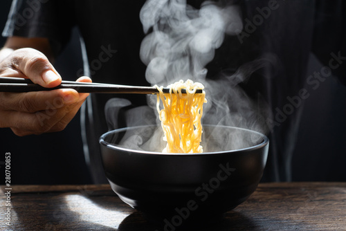 Hand uses chopsticks to pickup tasty noodles with steam and smoke in bowl on wooden background, selective focus. Asian meal on a table, hot food and junk food concept