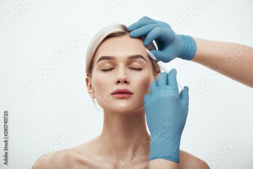 Cosmetic injection. Portrait of young pretty woman keeping eyes closed while doctors hand in blue medical gloves making an injection of botox in her face