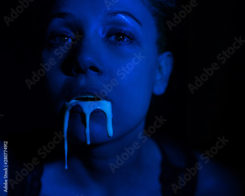 A thick white liquid flows down the lips of a young European girl. Dripping down. Ultraviolet, blue light. Close-up portrait. Spit out. 