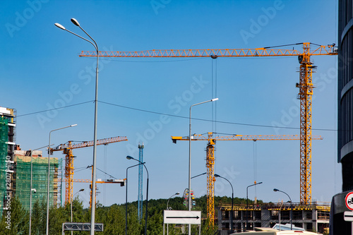 Several Yellow hoisting cranes against the blue sky. The cranes are lifting the load to the top of the house construction.