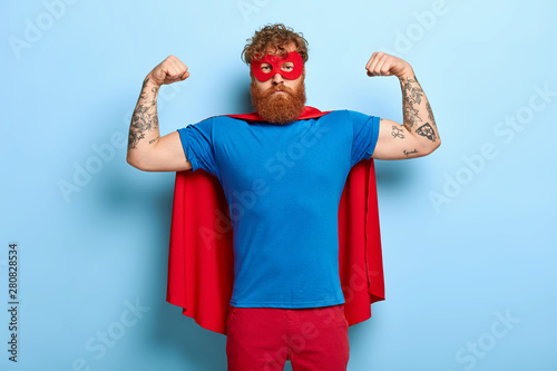 Successful hero wears red mask and cape, raises arms, shows biceps, demonstrates courage and strength, looks serious and confident, poses against blue wall. Real superhero ready to help you.