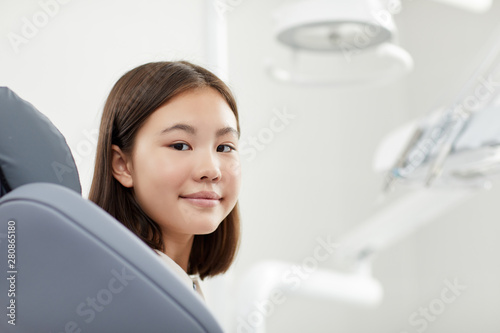 Portrait of teenage Asian girl sitting in dental chair in modern clinic and smiling at camera , copy space