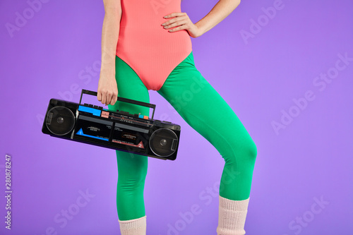 fitness girl stands in comfortable pink bodysuit and green leggins with one hand holding portable retro audio player and other is on her waist, has nice body curves and well-shaped
