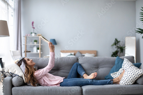 Beautiful smiling woman reading a book and lying on the sofa in the living room.