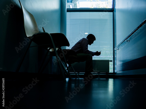 Low angle view of lonely patient in modern hospital waiting lobby room using smartphone mobile telephone as he waits for good or bad news from his doctor