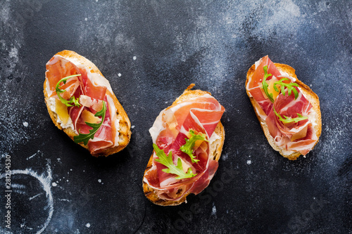 Open sandwiches with jamon, arugula and hard cheese on a concrete old dark background. Rustik style.Top view.