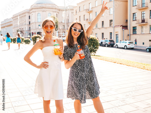 Two young beautiful smiling hipster girls in trendy summer clothes,dress.Sexy carefree women posing in the street.Positive models having fun in sunglasses.Drinking fresh cocktail smoozy drink