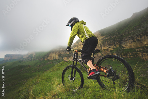 Aged male athlete in helmet and mask rides a mountain bike on a grassy slope against the background of plateau rocks and low clouds on an overcast day. Downhill mountain bike concept