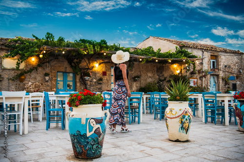 Young romantic woman walks near the colorful outdoor cafe in the beautiful sicilian coastal village Marzamemi in Sicily, south Italy