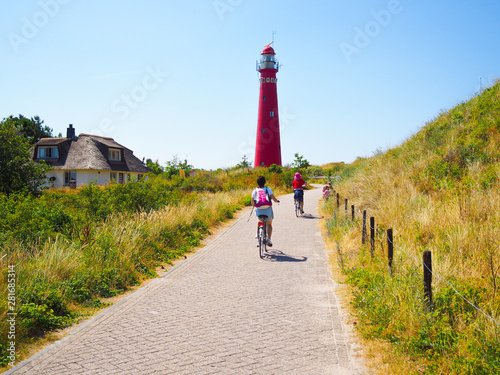 Back view cyclists traveling on the road heading towards to lighthouse in Schiermonnikoog islands, on sand dune against blue sky. Holland. Netherlands. Summer travel and vacation concept