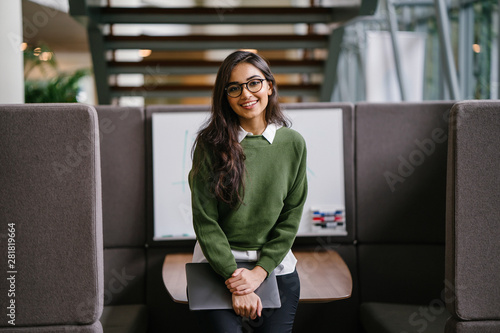 Portrait of a young, beautiful, intelligent and attractive Indian Asian MBA student smiling as she leans in a discussion booth in her campus. She is wearing a preppy outfit and smiling confidently.