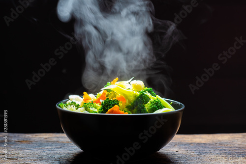 The steam from the vegetables carrot broccoli Cauliflower in a black bowl, a steaming. Boiled hot Healthy food on table on black background,hot food and healthy meal concept