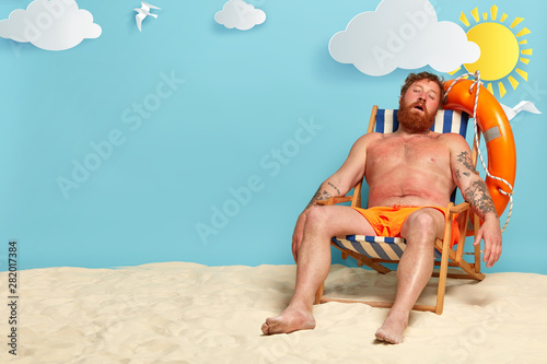 Relaxed bearded man sleeps on his deck chair, poses on beach, has red sunburned skin, sunbathes and tans, has summer vacation and holiday travel, beautiful sea view in background. People and rest
