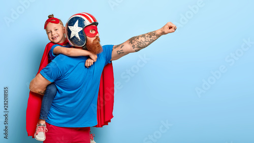Indoor shot of powerful dad in protective helmet carries small child on back, play superhero game, fly together and make good things for people, isolated on blue background. Childhood, parents concept