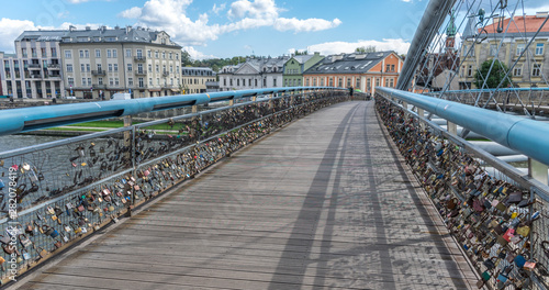 Horizontal perspective of the numerous padlocks in the Kładka Ojca Bernatka bridge in Poland, Krakow, with two blurred people walking in the background near an orange building.