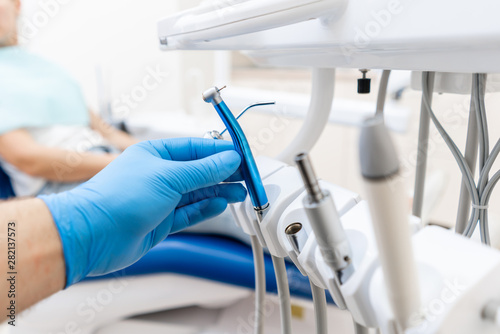 Close-up hand of dentist in the glove holds dental high speed turbine. The patient in blue chair at the background. Office where dentist conducts inspection and concludes.