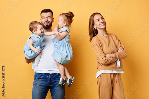 best help from husband, unforgettable Mother's day.unhappy depressed male babysitter cannot cope with his duty. carefree wife with crossed arms looking away. isolated yellow background, studio shot