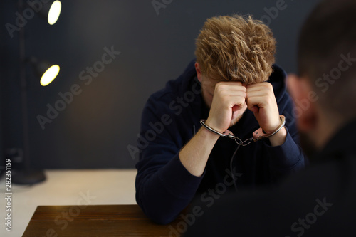 Police officer interrogating criminal in handcuffs at desk indoors, space for text