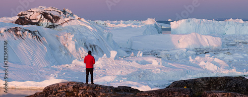 Travel wanderlust adventure in Arctic landscape nature with icebergs - tourist person looking at view of Greenland icefjord - aerial photo. Man by ice and iceberg, Ilulissat Icefjord.