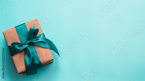 Gift box in craft wrapping paper and green satin ribbon on turquoise blue background, copy space right. Beautiful Christmas, New Year or Birthday present, flat lay or top view. Banner