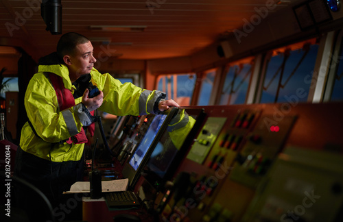 Navigator. pilot, captain as pat of ship crew performing daily duties with VHF radio, binoculars on board of modern ship with high quality navigation equipment on the bridge on sunrise.